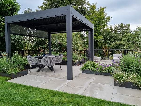 Dark grey metal pergola with adjustable textilene side partially lowered, over chairs on porcelain paved area. 