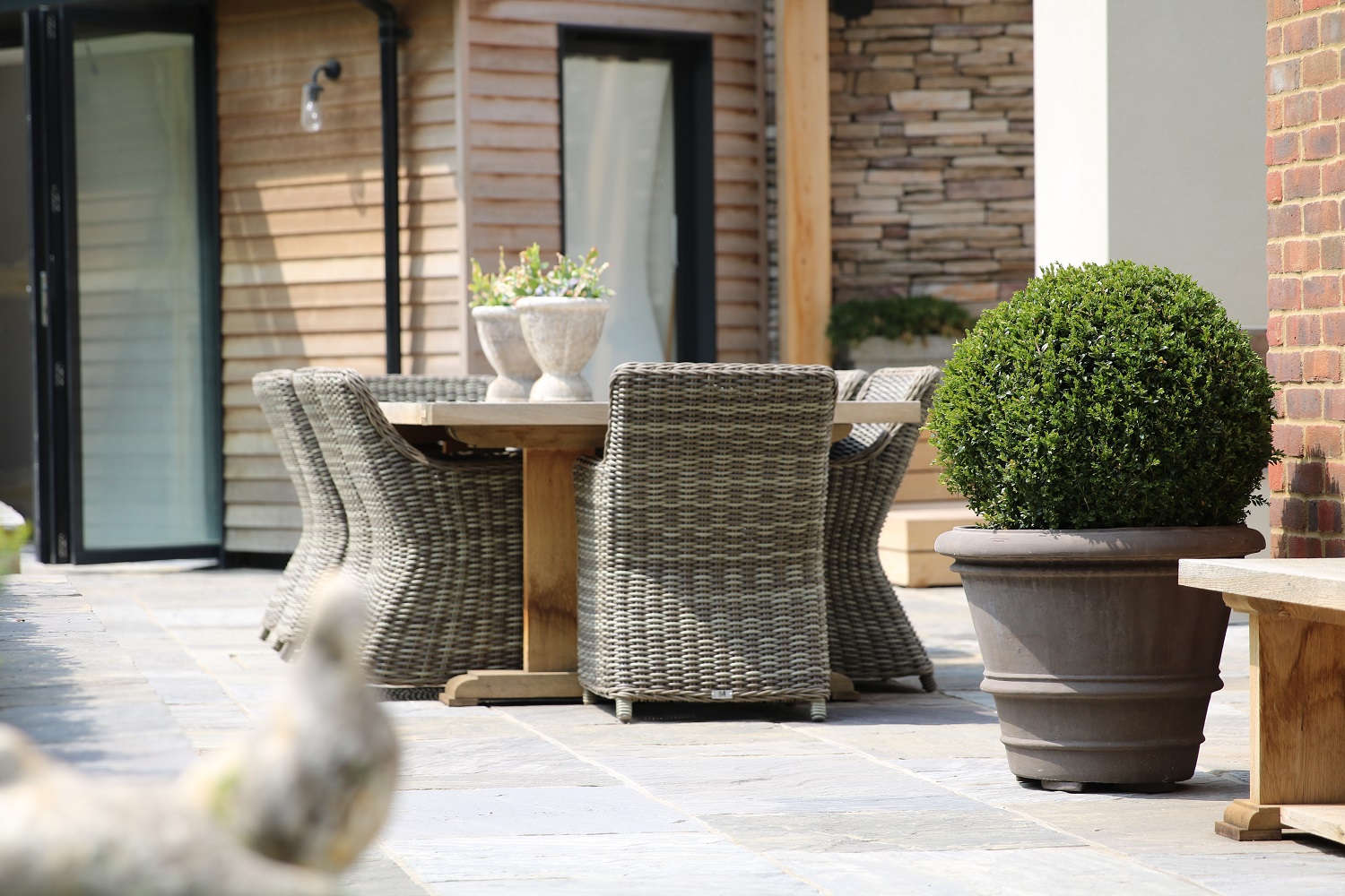 Trimmed shrub in pot on Black tumbled Indian sandstone paving with outdoor furniture with french doors in background.