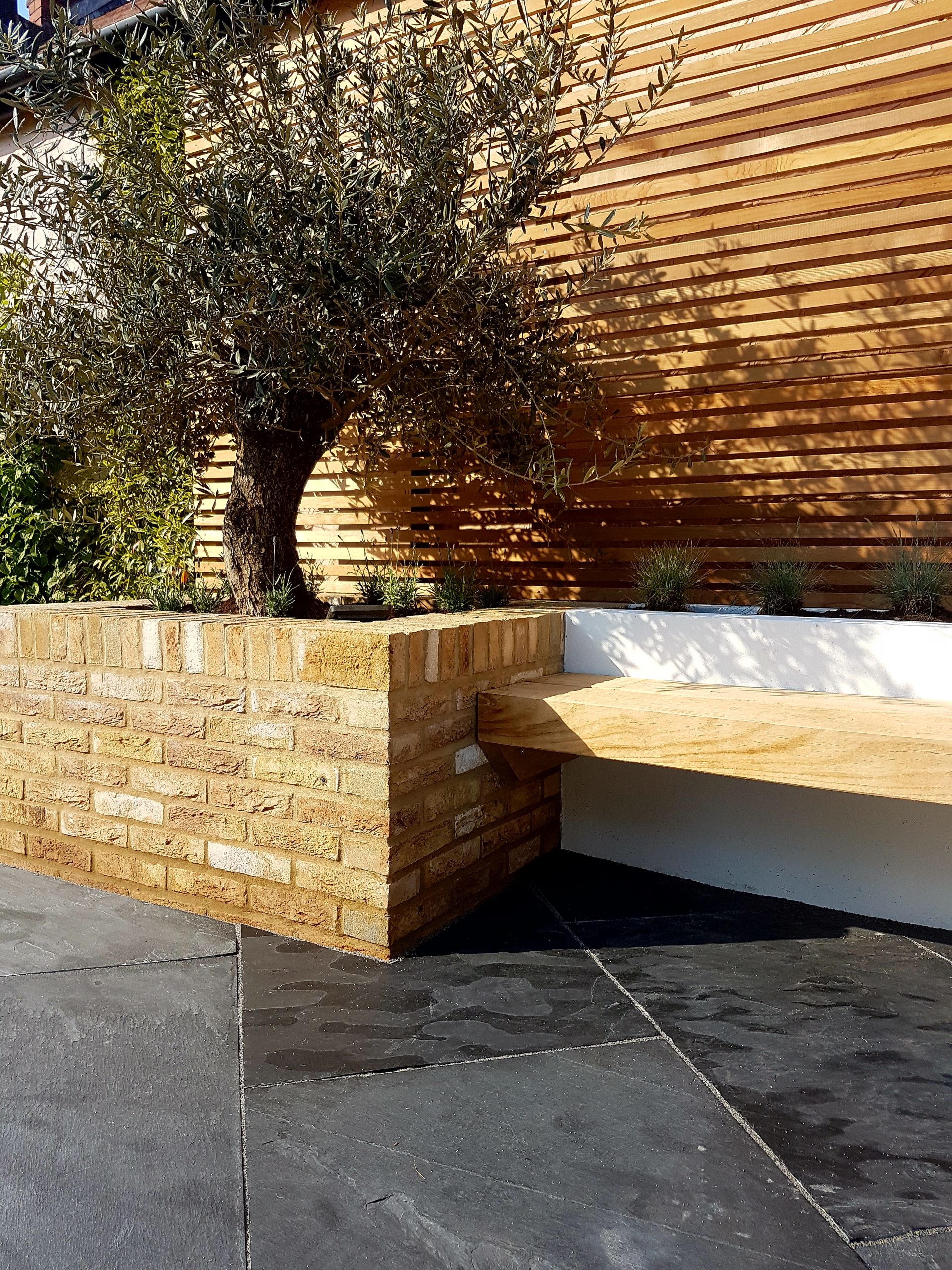 Cantilevered bench on rendered wall next to raised tree pit faced with outdoor wall cladding of facing bricks.