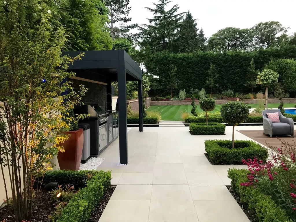 Outdoor kitchen under narrow pergola on large patio with topiary and furniture. 