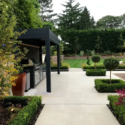 Outdoor kitchen under narrow pergola on large patio with topiary and furniture.