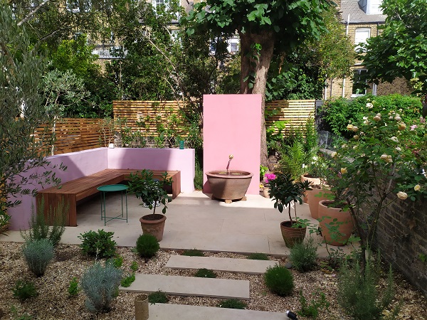 Small garden with L-shaped raised beds with bench, Golden Stone porcelain paving and water feature. 
