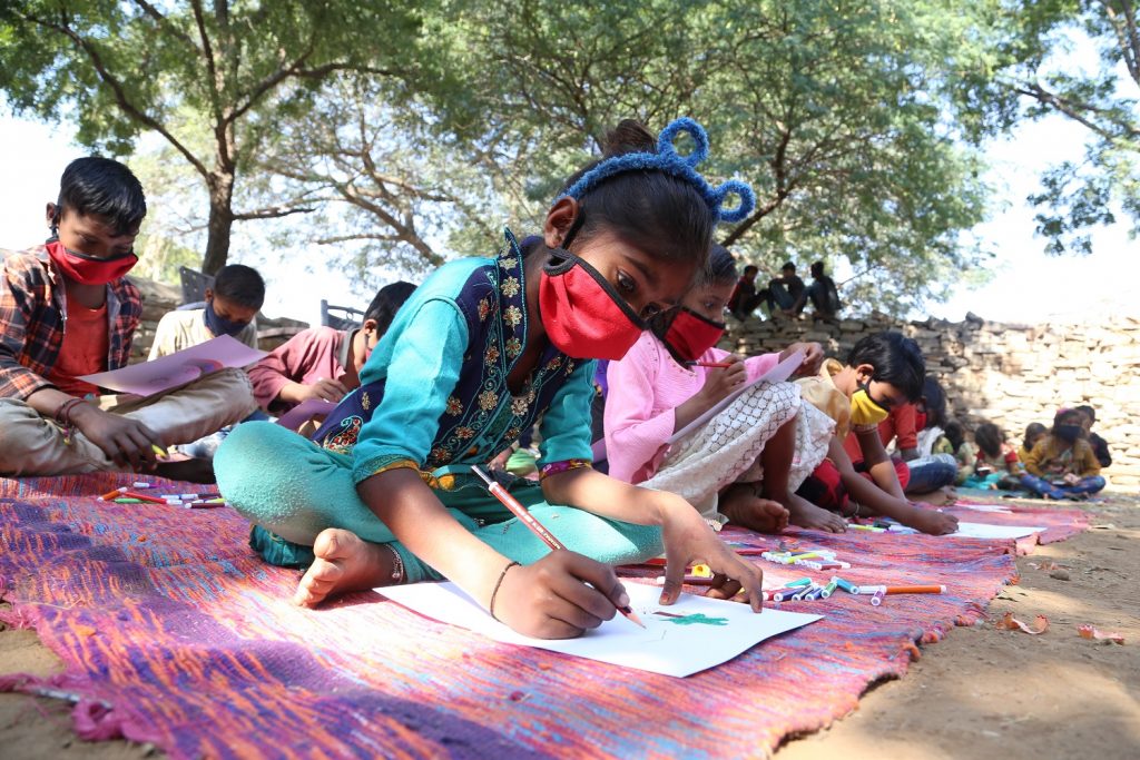 A class of young children sitting on the ground in open air class, wearing masks, Rajasthan, India, No Child Left Behind