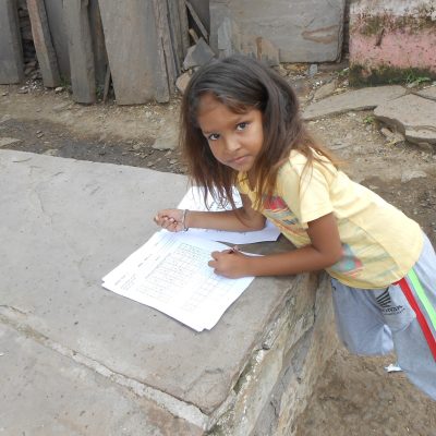 Young girl leaning over, solving worksheets, resting on pile of stone slabs, in Budhpura, Rajasthan, India