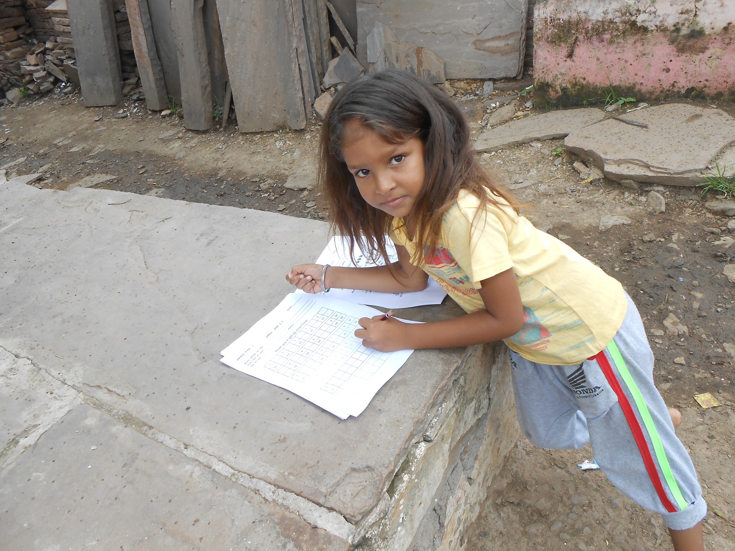 Young girl leaning over, solving worksheets, resting on pile of stone slabs, in Budhpura, Rajasthan, India