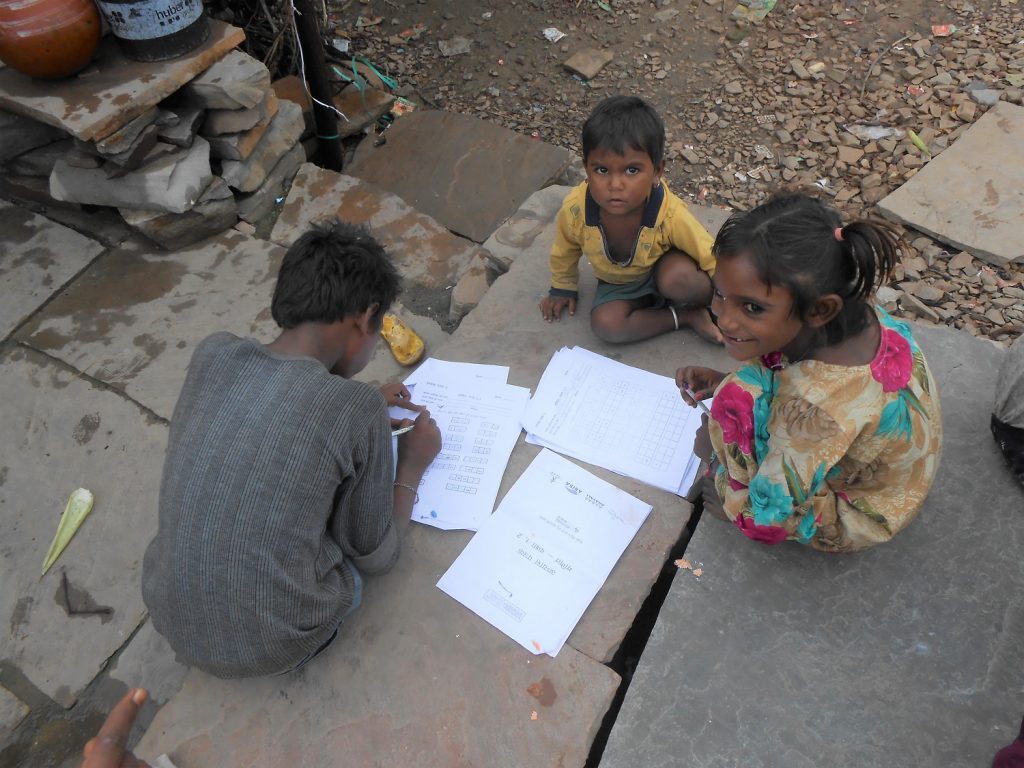 Group of 3 children of different ages, sitting on stone slabs, with worksheets spread around. in Budhpura, Rajasthan, India, No Child Left Behind