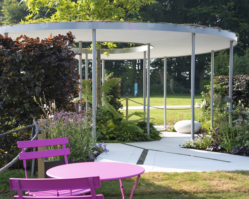 The Cancer Research Legacy Garden, RHS Tatton Park 2021, pink bisto set in foreground, white doughnut shaped roof with tree growing through hole, white paving with rills, designed Neil Sutcliffe, built Creative Roots, Florence White Porcelain, London Stone