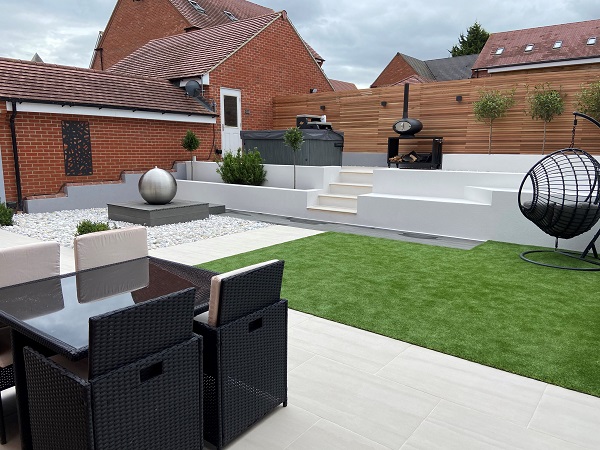 Raised area terraced in Faro porcelain paving and coping runs down length of fence with hot tub and pizza oven.