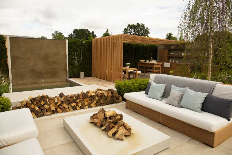 Natural stone patio idea of Fire table of single slab of Jura Beige sits between cushioned benches in  RHS Hampton Court 2021.