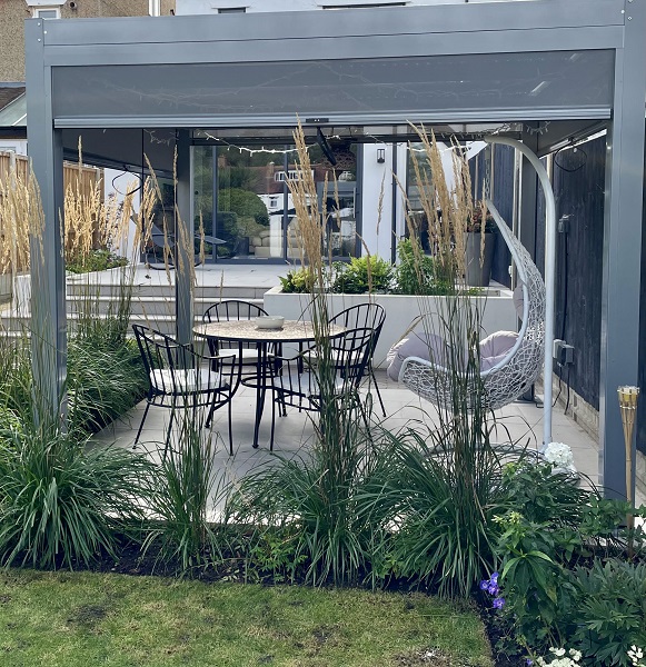 Aluminium pergola with sides over patio with round table and chairs. Wide steps up to house in background. 