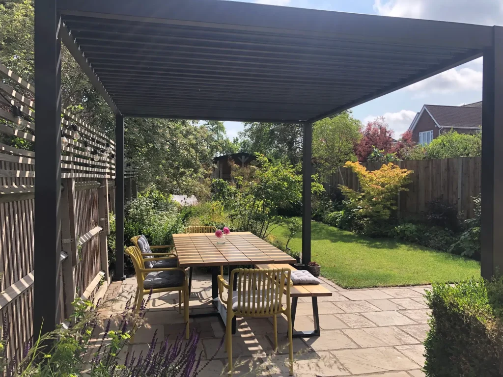 Grey Proteus modern metal pergola over dining set on patio by fence and on edge of rectangular lawn.