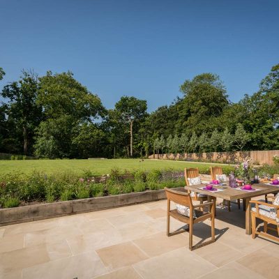 Sealed Harvest Sawn Sandstone patio with with wooden dining table and chairs with large lawn behind.