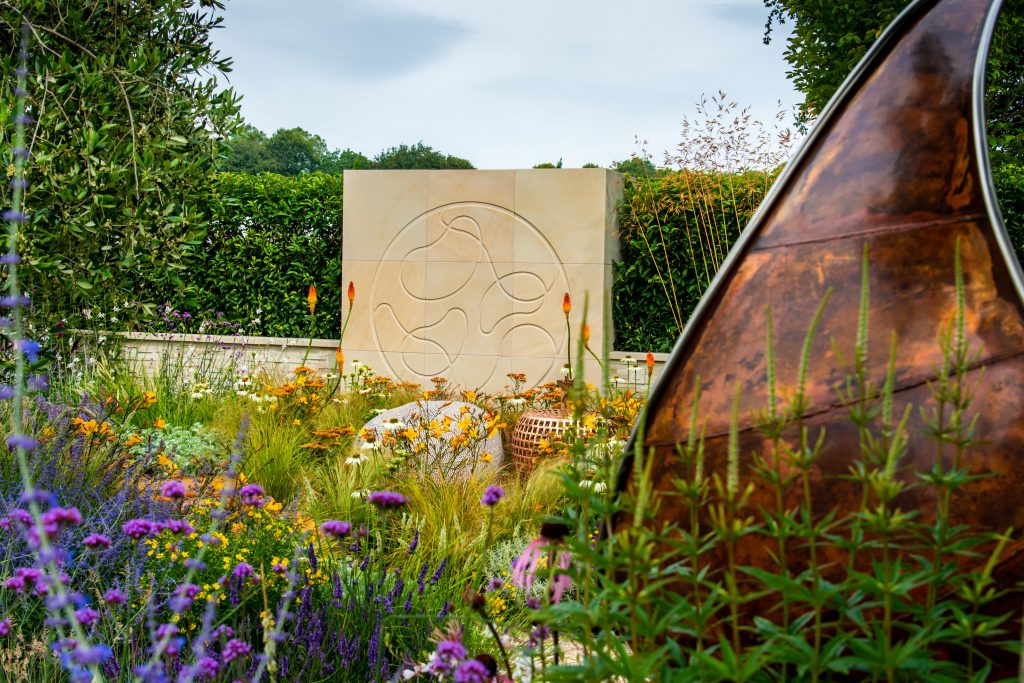 Harvest Sandstone feature wall with engraving with hedges behind, planting in front and metal sculpture. 