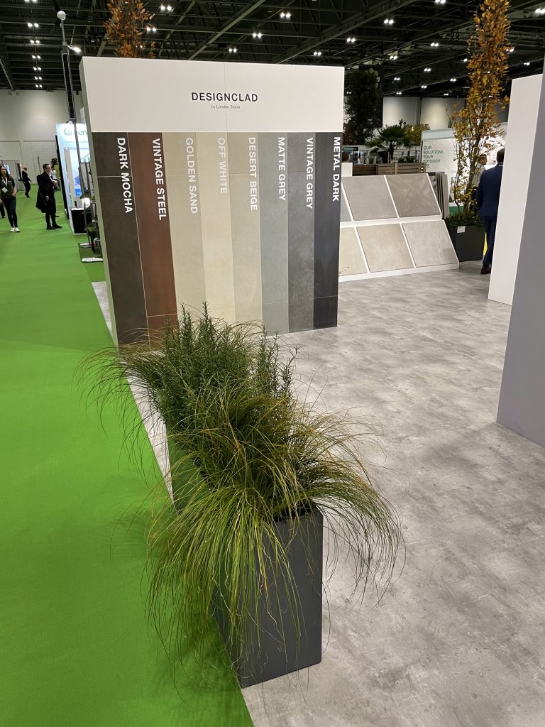 A planter filled with plants sits in the foreground. DesignClad and 800x800 porcelain paving samples can be seen in the background.