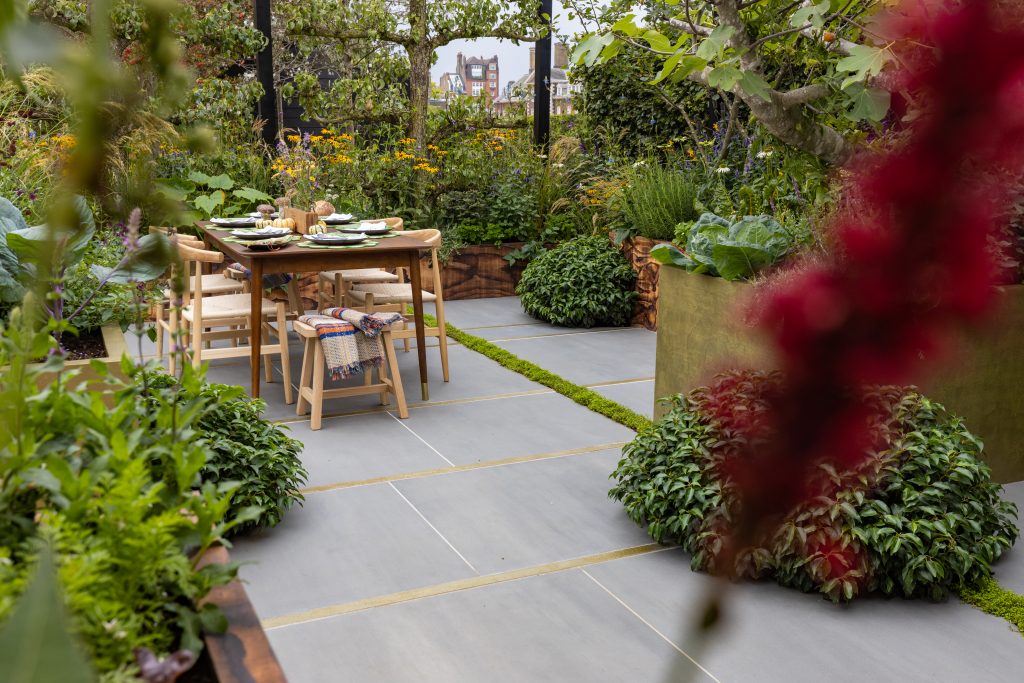 Sidewalk Porcelain is laid with a wooden outdoor table and chairs set sitting on top and surrounded by full plant plots.