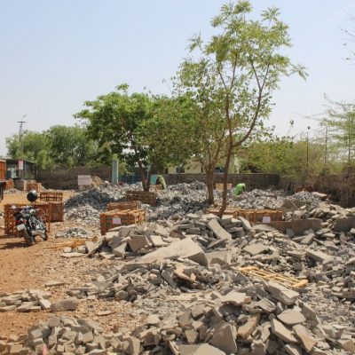 Cobble yard in Budhpura, India, with piles of stone under small trees, lorry on left, and women in distance