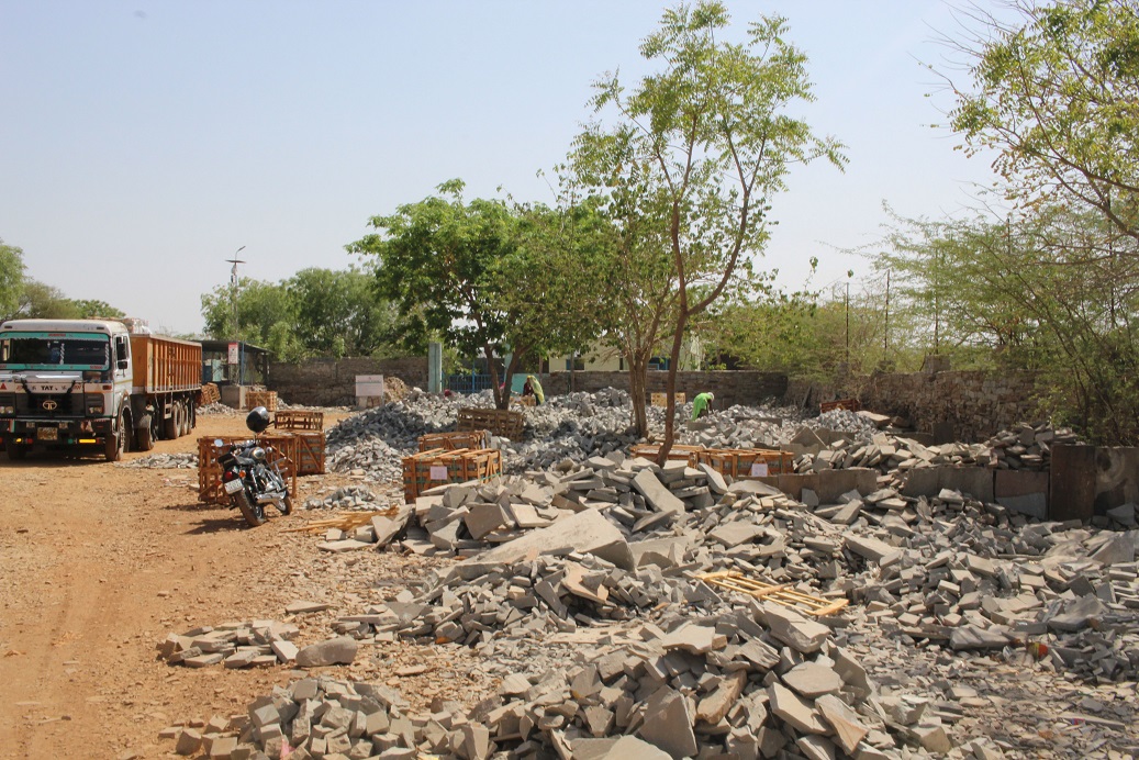 Cobble yard in Budhpura, India, with piles of stone under small trees, lorry on left, and women in distance