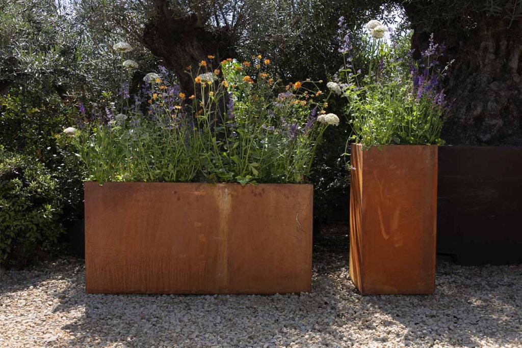 Corten Steel Tall Trough Planted and Corten Steel Tall Cube Planter stood side by side, filled with orange, purple and white flowers. 