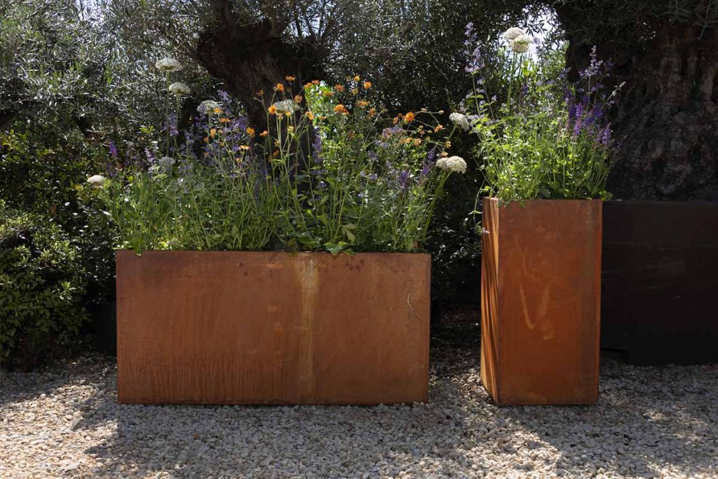 Corten steel tall square planter and trough planter stand next to each other on gravel. Both planted. 