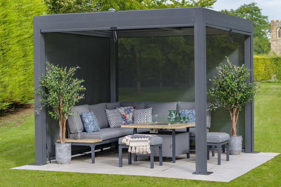 A metal pergola is positioned above a grey modular sofa dining set on a small paved square, in the center of a lawn.