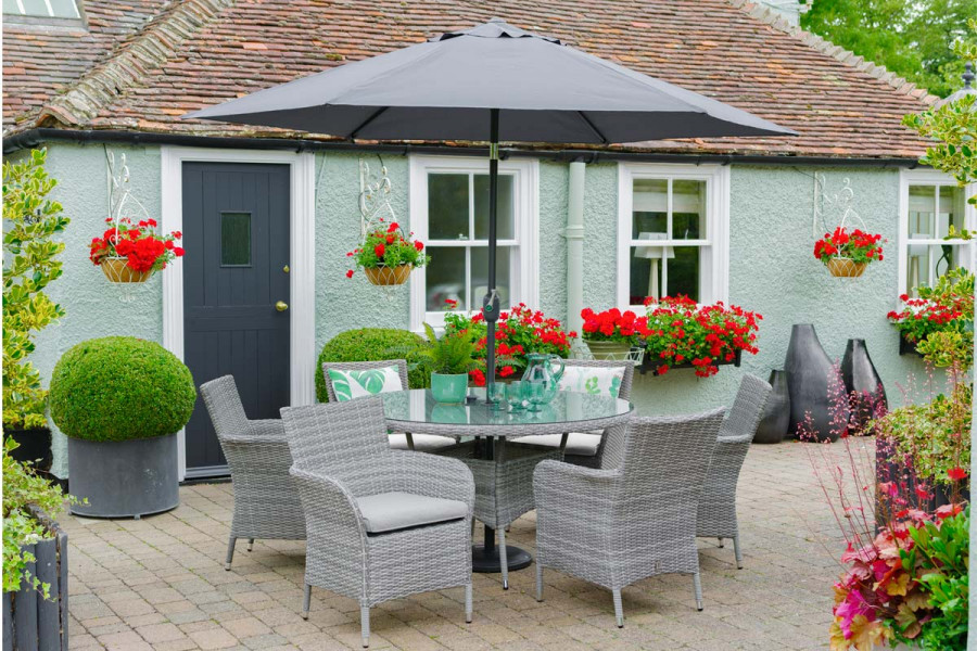 6 seat circular rattan dining set and parasol in a charming garden setting with vibrant red planting. 
