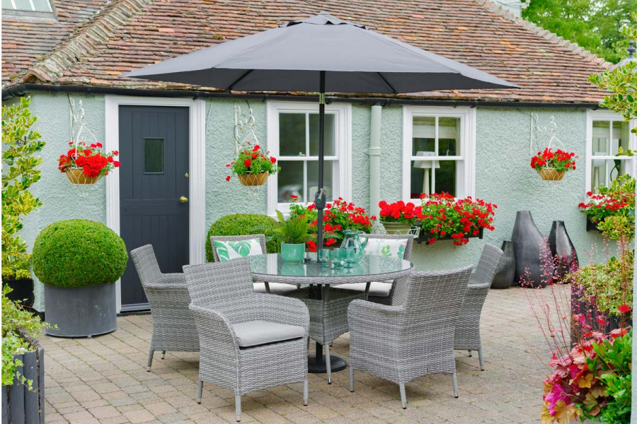 6 seat circular rattan dining set and parasol in a charming garden setting with vibrant red planting.
