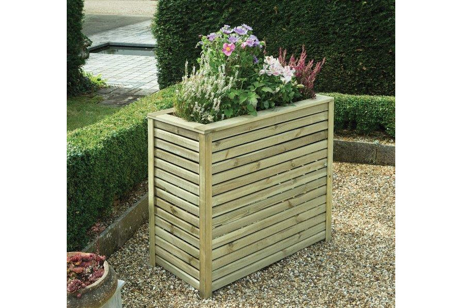 Slatted tall trough planter of pressure-treated redwood, planted and sitting on gravel by low hedge.