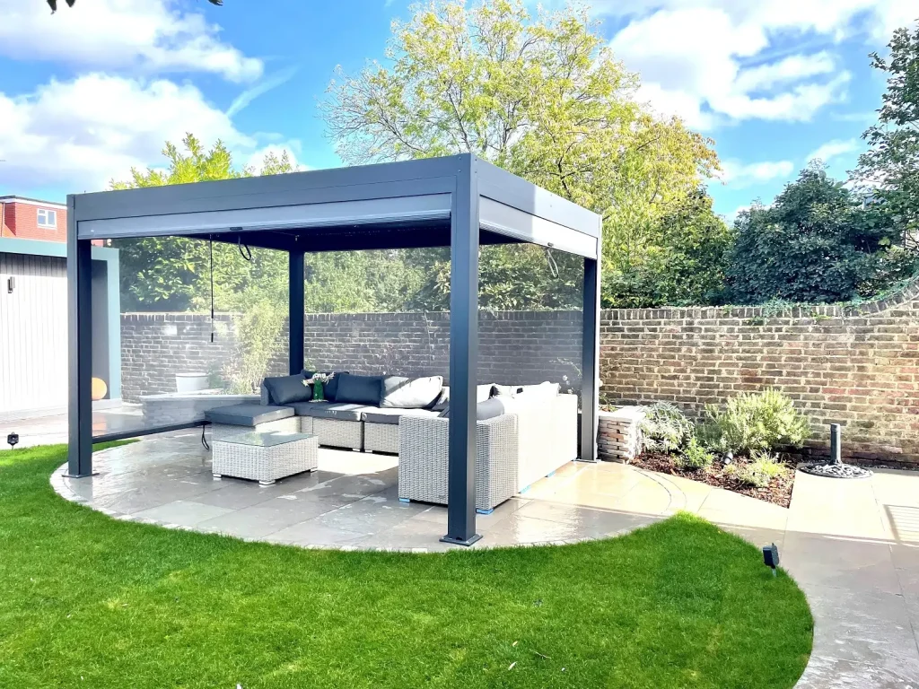 Dark grey metal pergola, 2 sides lowered, over oval patio with curved path leading away.,