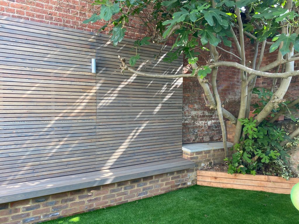 1800cm larch slatted fence panel affixed to tall brick wall with bench seat below.