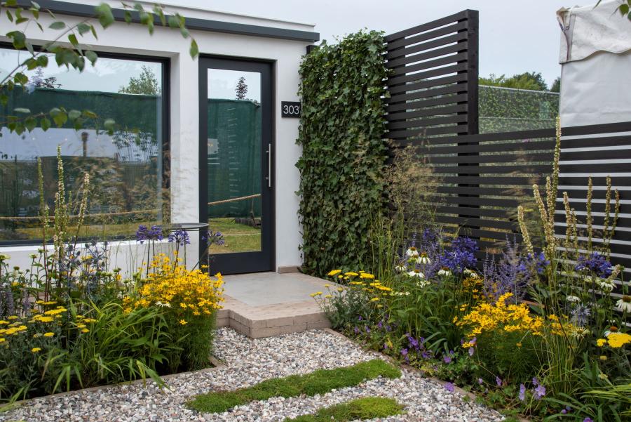 Fence of open slatted Dark Ash composite battens behind planted border in front of front door of single storey building.