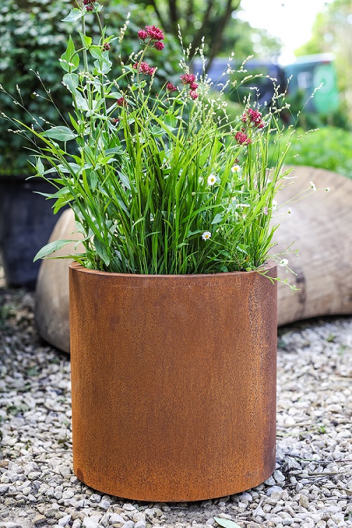 Round corten steel planter filled with grasses, sitting on gravel in front of large log.