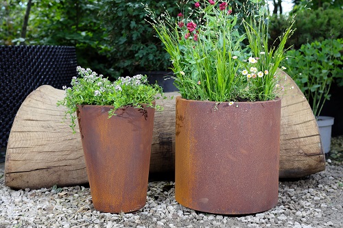 Tapered and Round Corten Steel Planter filled with plants sit on gravel in front of large log.