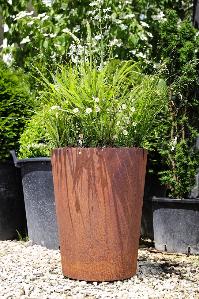 400 x 500mm tapered Corten steel planter sits on gravel in front of grey weathered wooden screen.