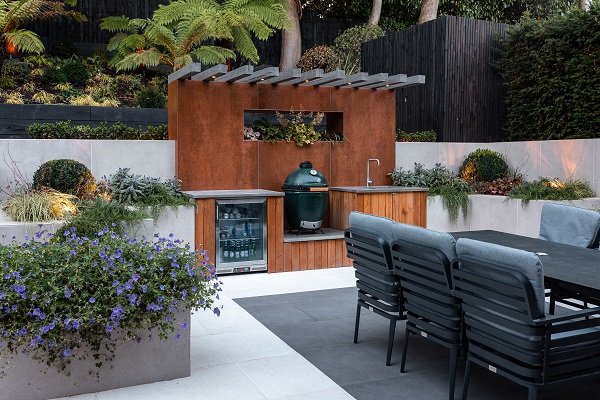 Outdoor kitchen with small canopy and porcelain tile splashback at back of seating area with planters and black and white paving.