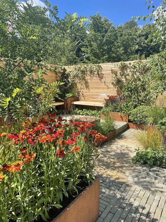 Larch fencing surrounds Lunch Break Garden, RHS Hampton Court 2022, with bench and red flowers.