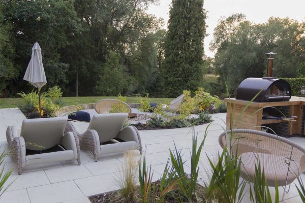 Sandy white porcelain paving for patios in design with inset beds, outdoor kitchen and sunloungers. Built by Creative Roots.