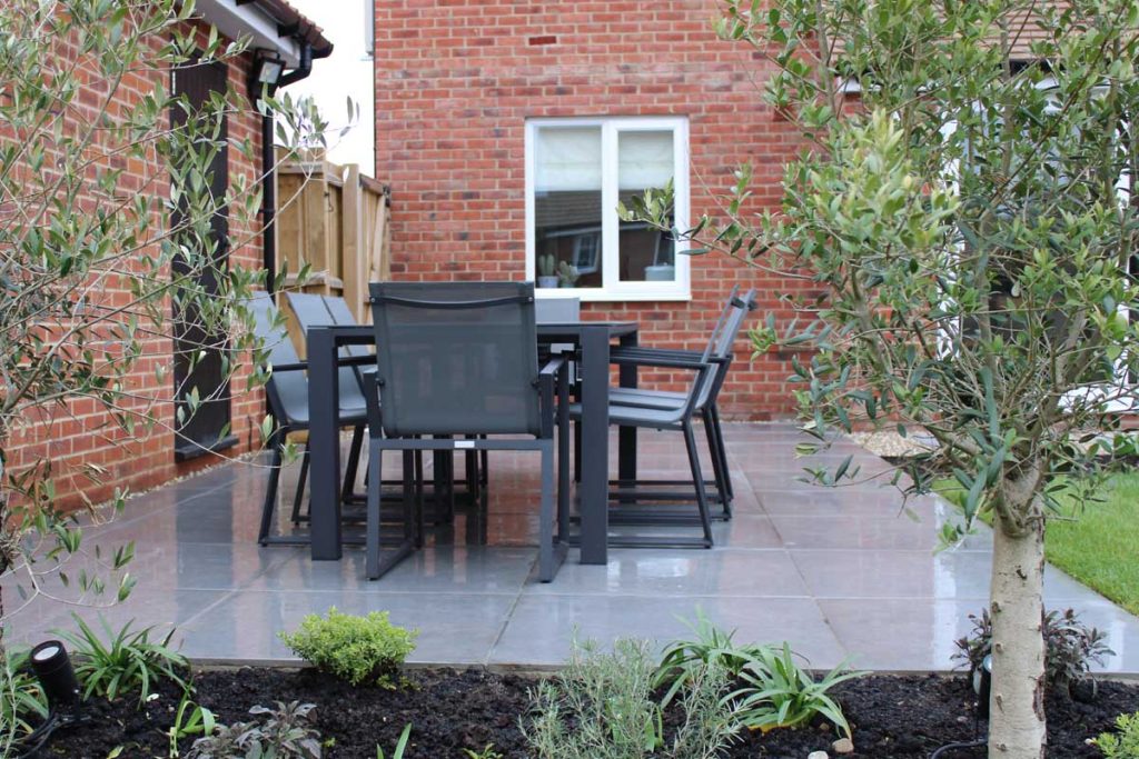 Metal dining set on anthracite porcelain 800x800 paving slabs with house, lawn and planted bed.