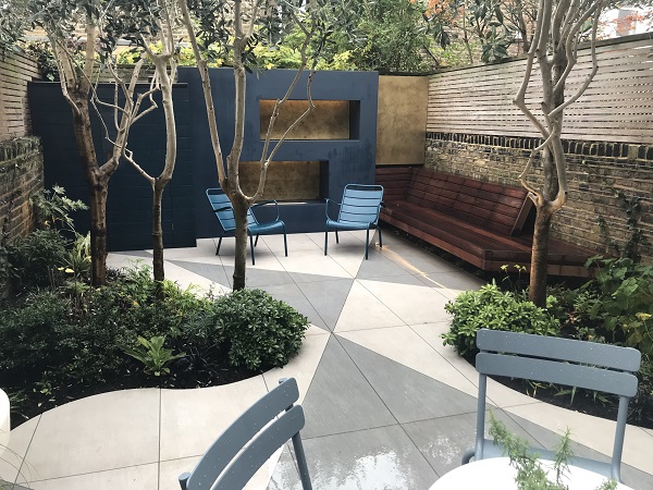 Urban back garden laid with bespoke porcelain paving for patios, with chairs, slatted fence and brick wall.