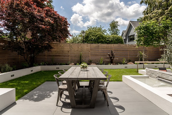 Wooden table and 6 chairs on patio of Sidewalk XL porcelain slabs, next to water feature and lawn.