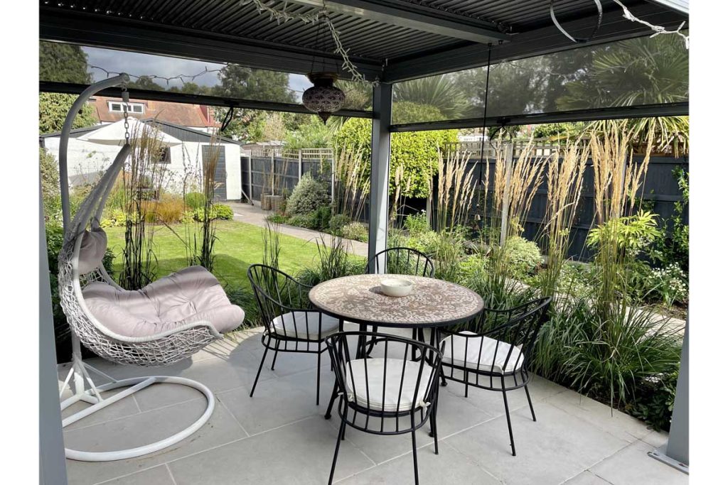 LG dark grey aluminium pergola over garden dining set, sides partially lowered.Design by Emily's Gardens, built by JR Landscaping