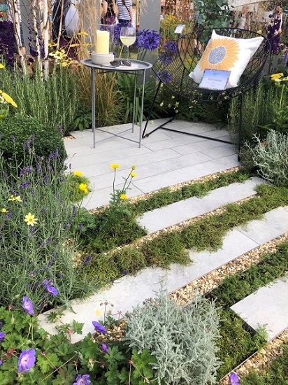 Light Grey porcelain planks surrounded dense planting and wire chair and table .