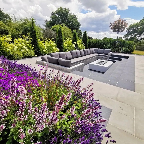A sunken seating area with long sofa and white firepit table is laid with Slab Coke porcelain in midst of paler paving.