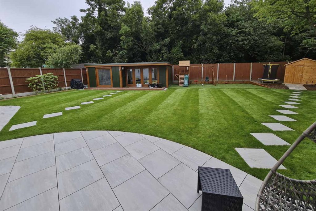 Large garden with patio in Kandla Grey Porcelain garden tiles and paths leading across lawn to garden buildings.