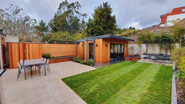 Right-angled patio of Ash Beige porcelain paving slabs with lawn and stained wooden garden room and fence. 