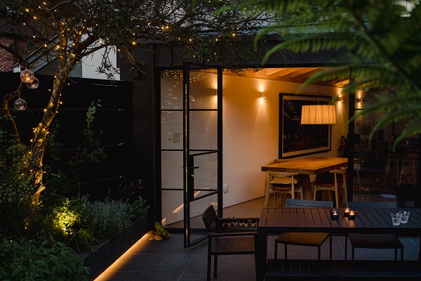 Illuminated interior with Crittall doors opening onto Basalt Porcelain patio with lights in trees and planted border.