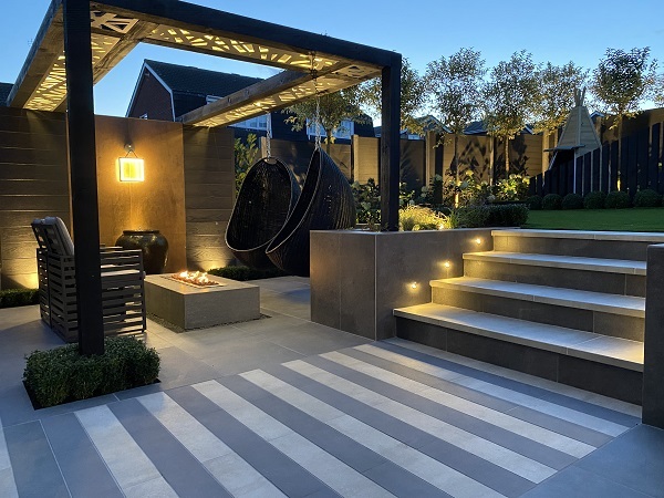 Seating area with garden lighting, firepit under pergola, cream and steel grey porcelain paving, steps up to lawn.