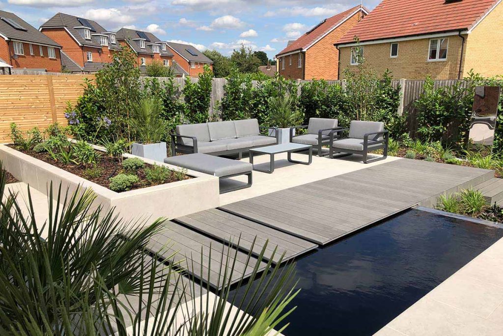 Charcoal composite decking partly covers square pond in front of paved area with modular furniture.