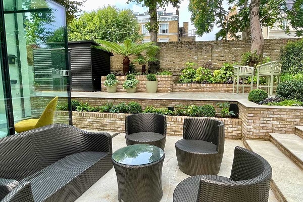 Rattan furniture sits on beige porcelain paving slabs edged with London brick raised beds.