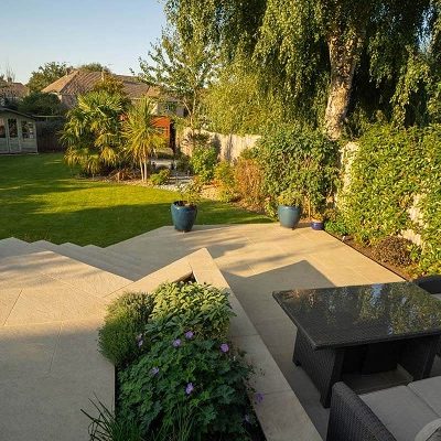 Beige porcelain patio with steps descends to lawn in large fenced garden with mature trees.