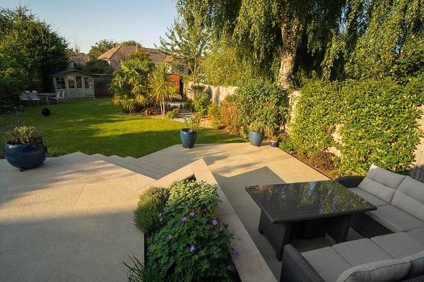 Beige porcelain patio with steps descends to lawn in large fenced garden with mature trees.